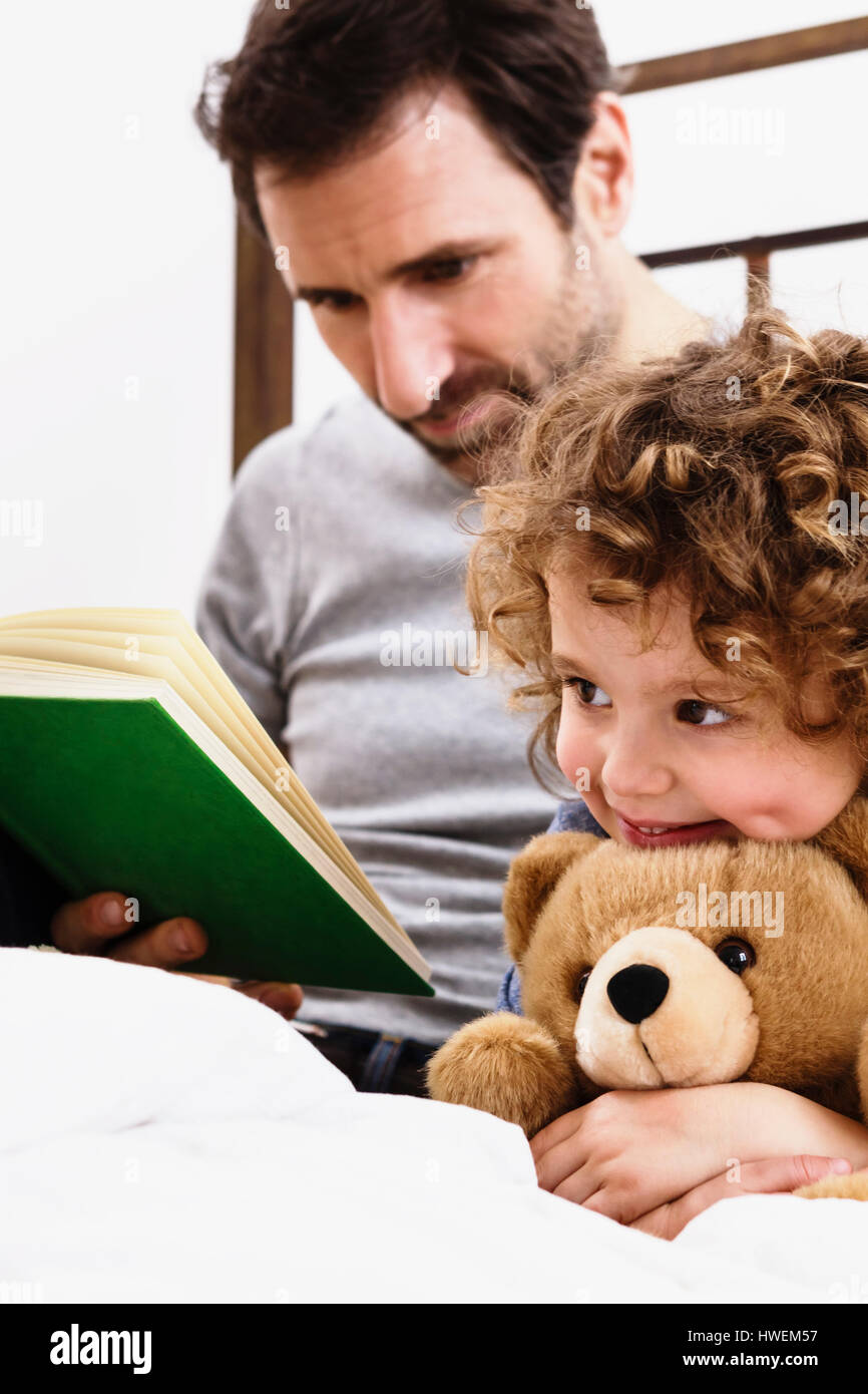 Girl hugging teddy bear while father reads storybook in bed Stock Photo