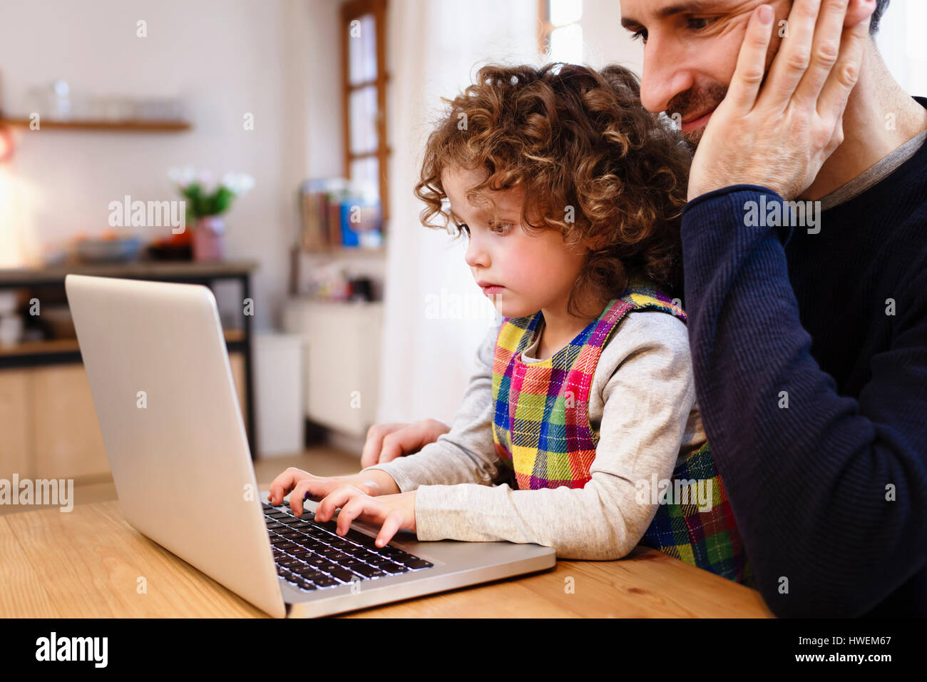 Girl on father's lap typing on laptop in kitchen Stock Photo