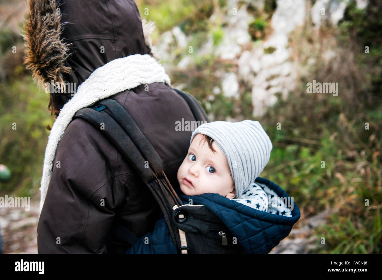Portrait of baby boy wearing knit hat, carried in baby sling by mother Stock Photo