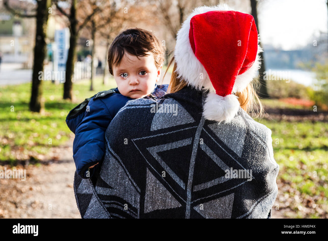 Over shoulder view of young woman in santa hat carrying baby boy in park Stock Photo