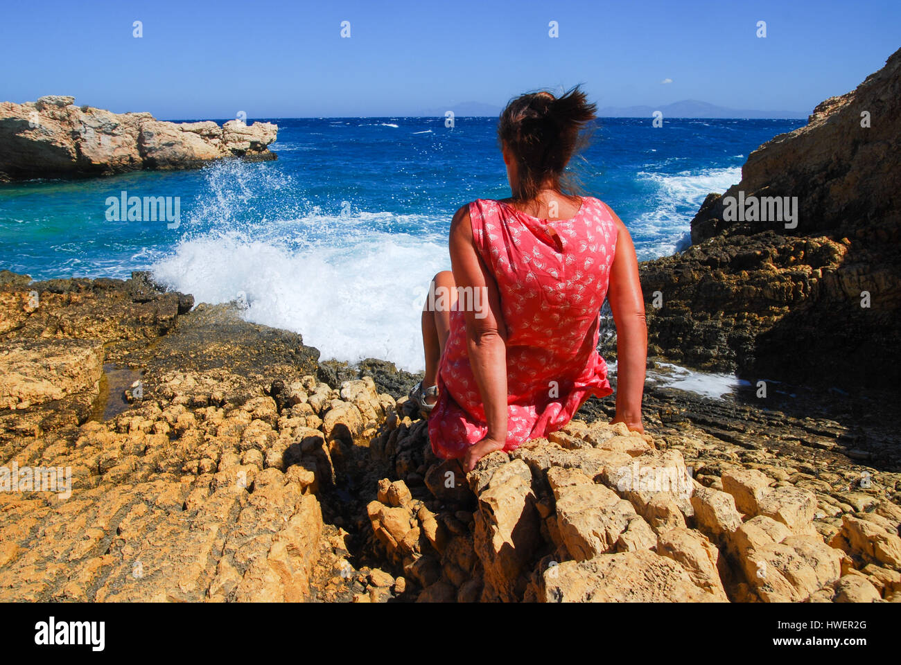Woman leaning back on rocks facing the sea as waves splash. Stock Photo