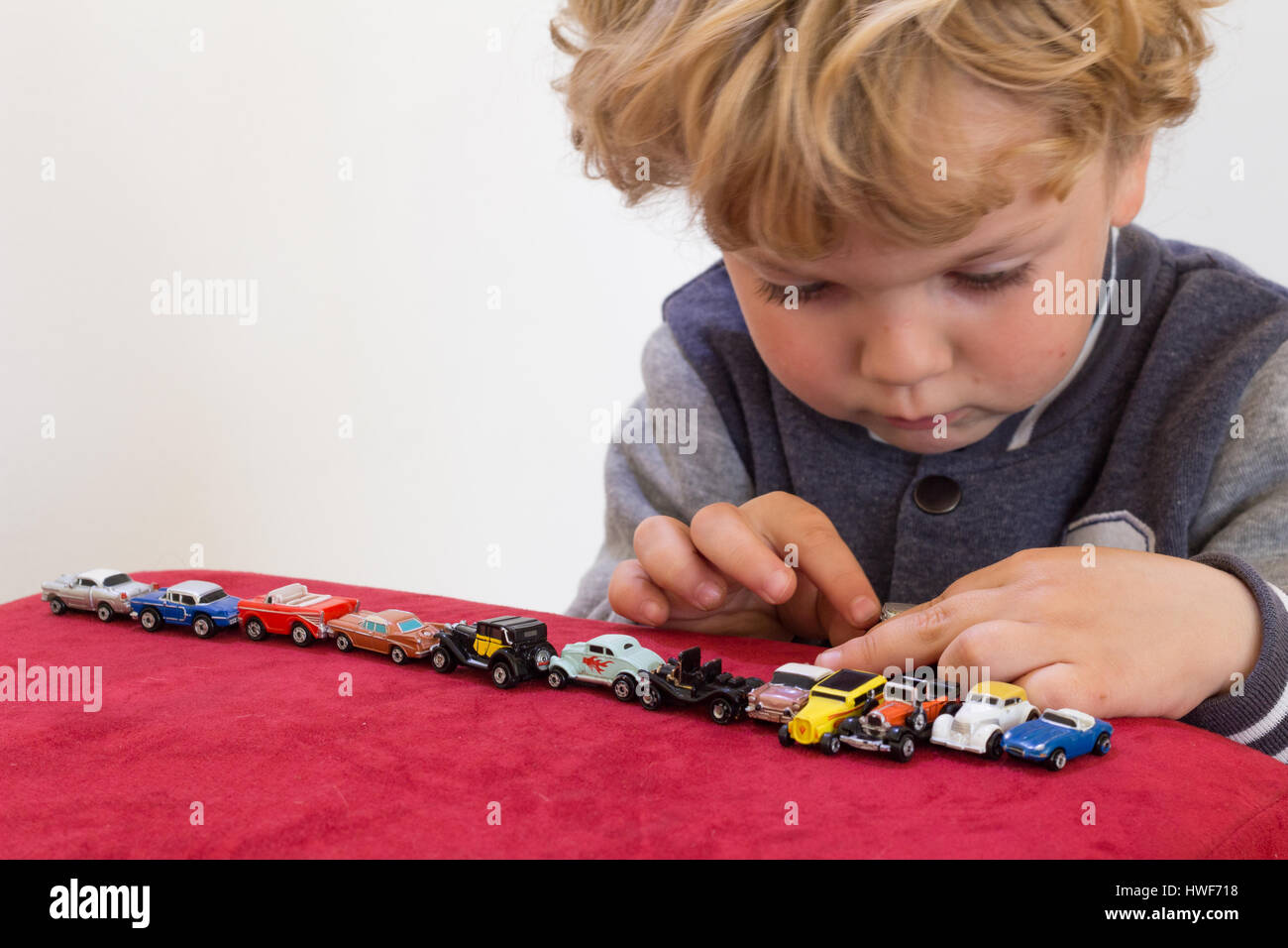 QUEENSTOWN, SOUTH AFRICA - MAY 17 2015 - Little blonde boy playing with vintage toy cars on red velvet pouf chair - Focus on his hands opening a tiny  Stock Photo
