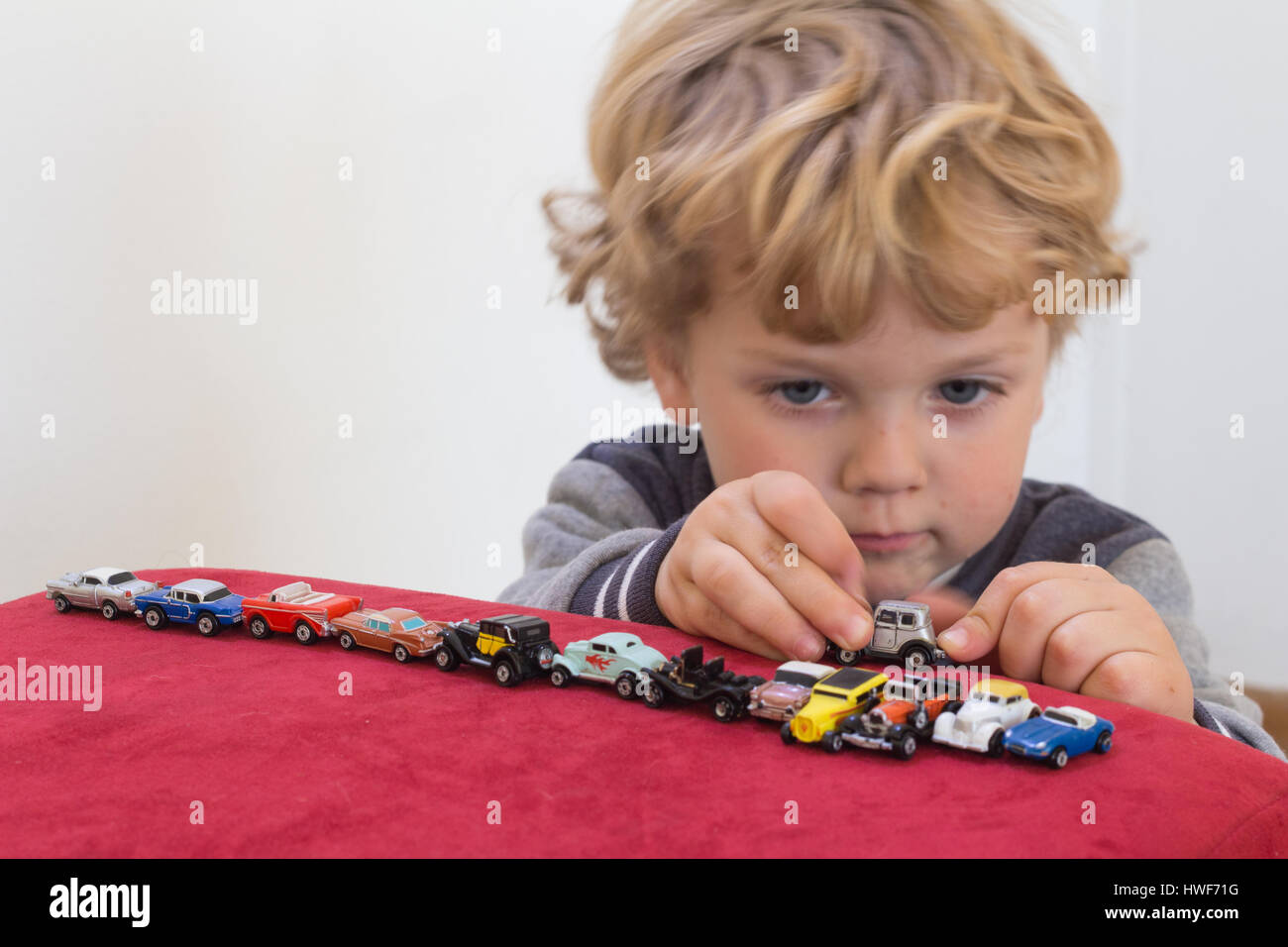 QUEENSTOWN, SOUTH AFRICA - MAY 17 2015 - Little blonde boy playing with vintage toy cars on red velvet pouf chair - Focus on his hands and little cars Stock Photo
