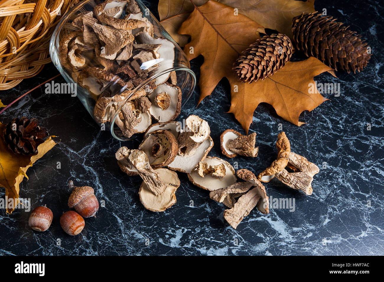 Dry white wild mushrooms in glass jar and on the table. Yellow wooden basket on back background, several dry oak leaves, acorns and fir cones. Composi Stock Photo