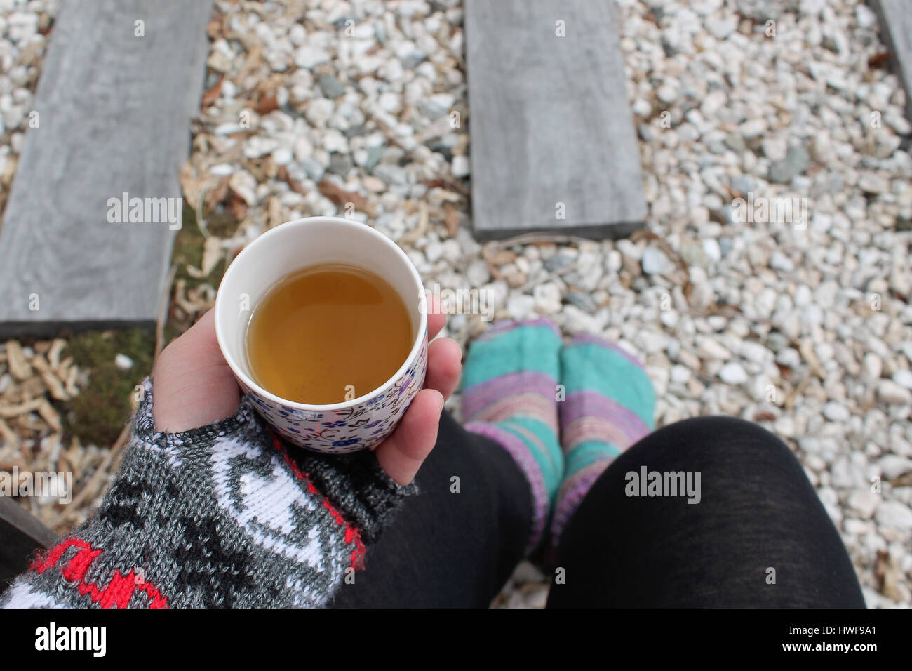 Top view of lady sitting down in muted grey outdoor light, holding a cup of tea, wearing cosy winter colourful socks and mittens. Stock Photo