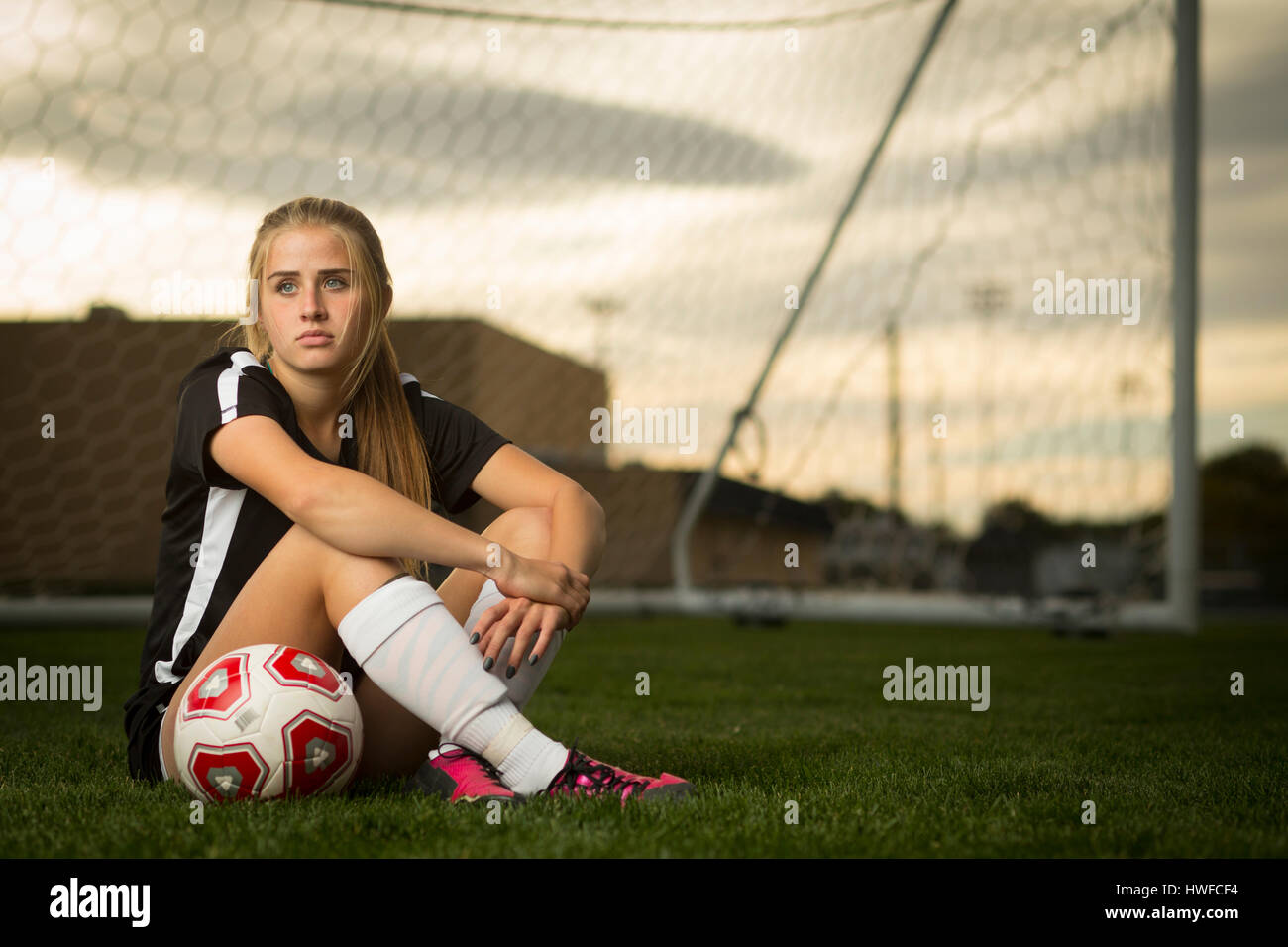 Athlete sitting with ball in soccer goal under sunset sky Stock Photo