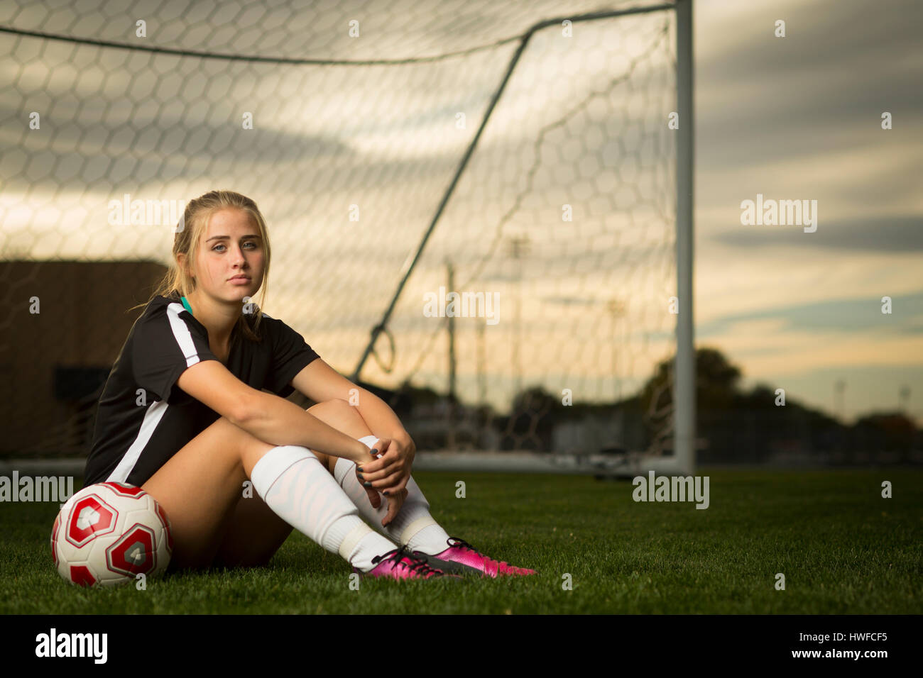 Athlete sitting with ball in soccer goal under sunset sky Stock Photo