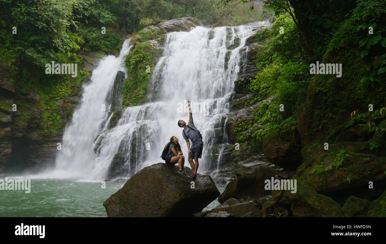 Low angle view of couple standing on boulder pointing and admiring scenic view near tropical waterfall Stock Photo