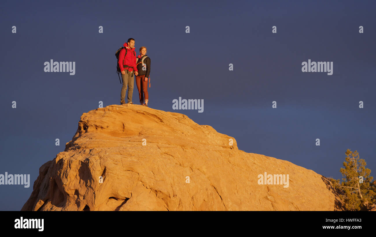 Low angle view of couple standing on remote boulder under sunset sky Stock Photo