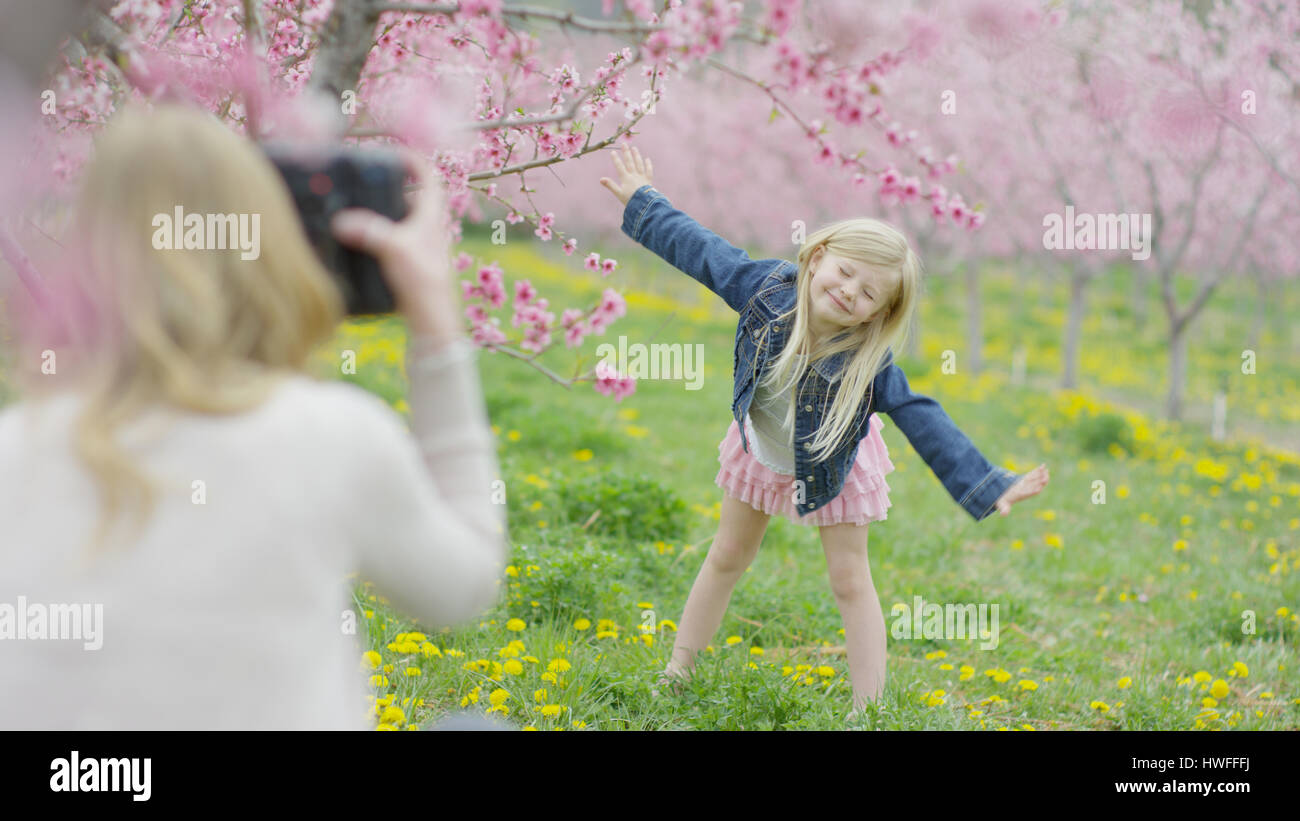 Mother photographing playful daughter posing in park field with flowering trees Stock Photo