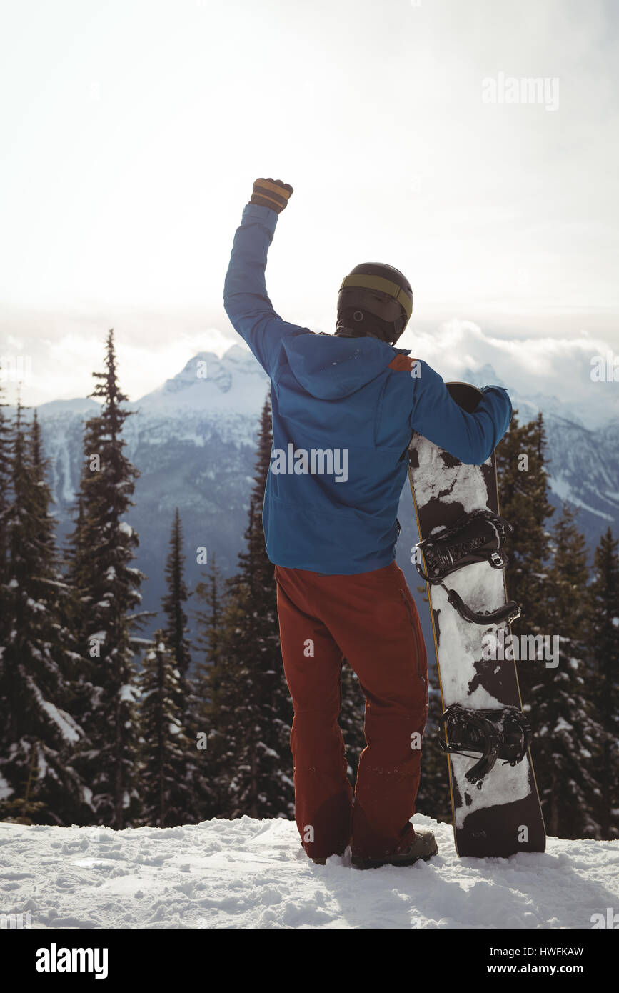 Rear view of man with hand raised holding snowboard on mountain against ...