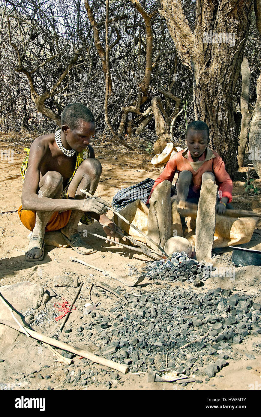 Local blacksmith of Datoga Tribe and his children working in the shade of an Acasia tree.  Lake Eyasi, northern Tanzania. Stock Photo