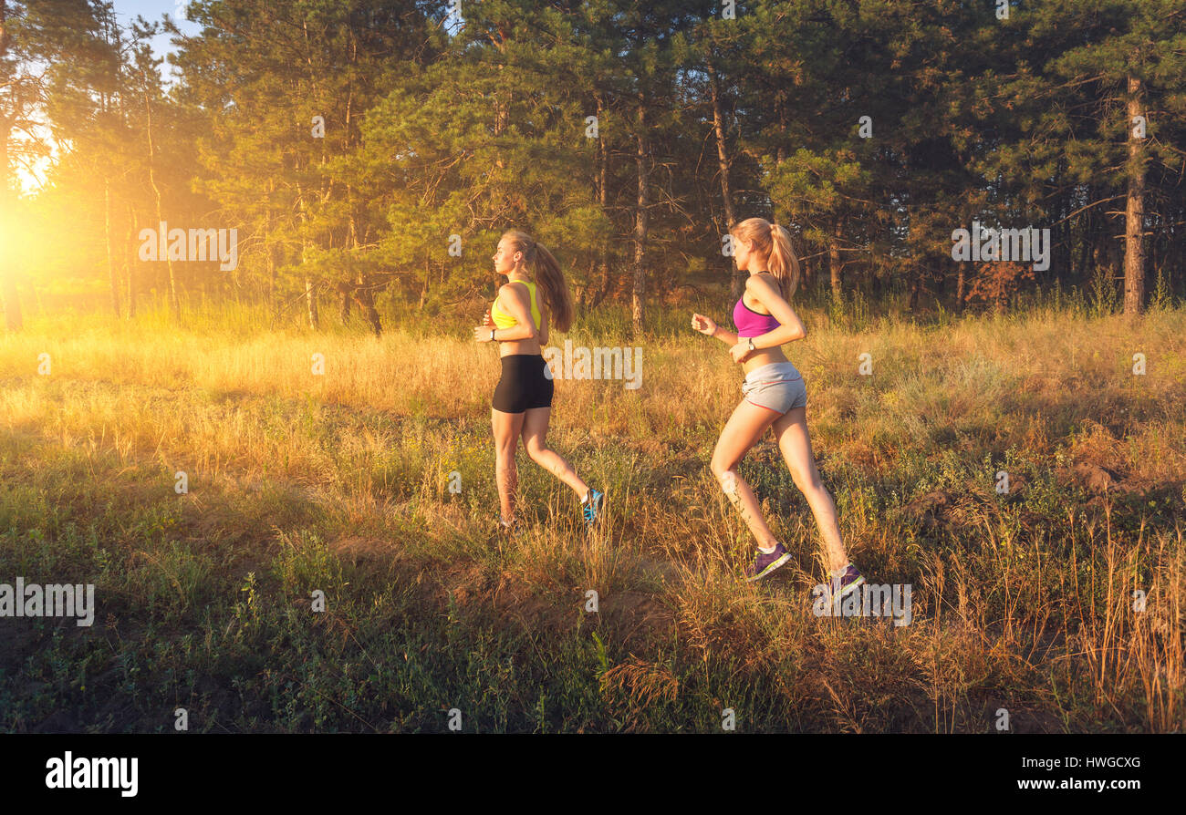 Young sporty girls running on a field near the trees at sunset in summer. Athlete running on the off road in the evening. Active woman. Sport and heal Stock Photo
