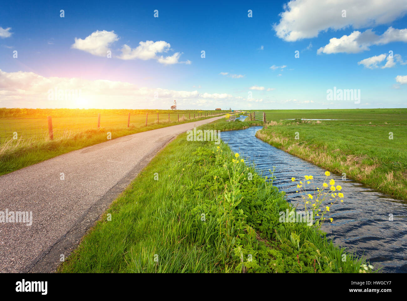Landscape with green grass field, road, lighthouse, river and blue sky with clouds reflected in water at amazing sunset in spring. Colorful nature bac Stock Photo