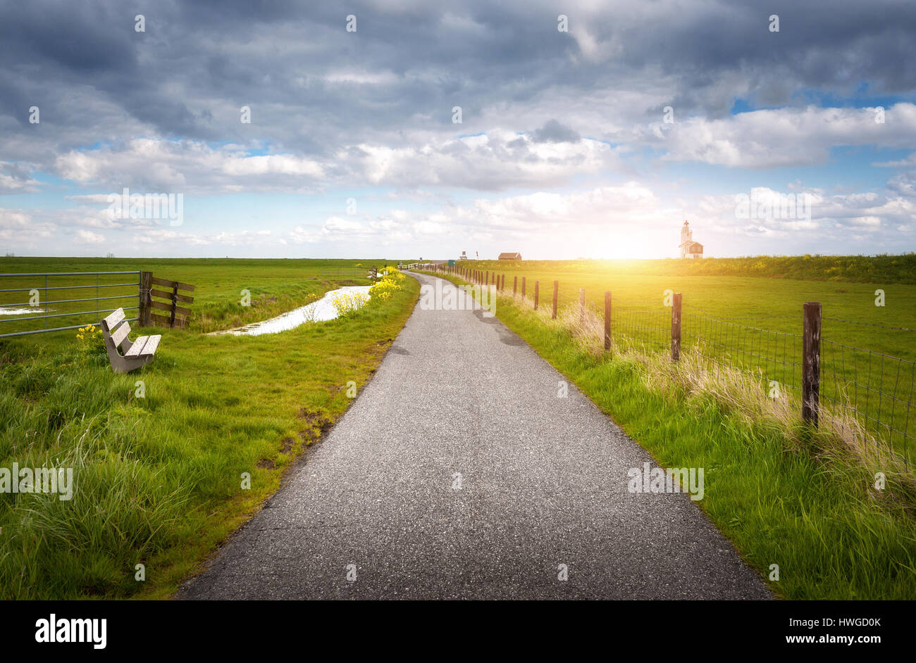 Beautiful rural road, colorful green grass, yellow flowers on the background of dramatic blue cloudy sky and lighthouse at sunset in spring. Amazing l Stock Photo