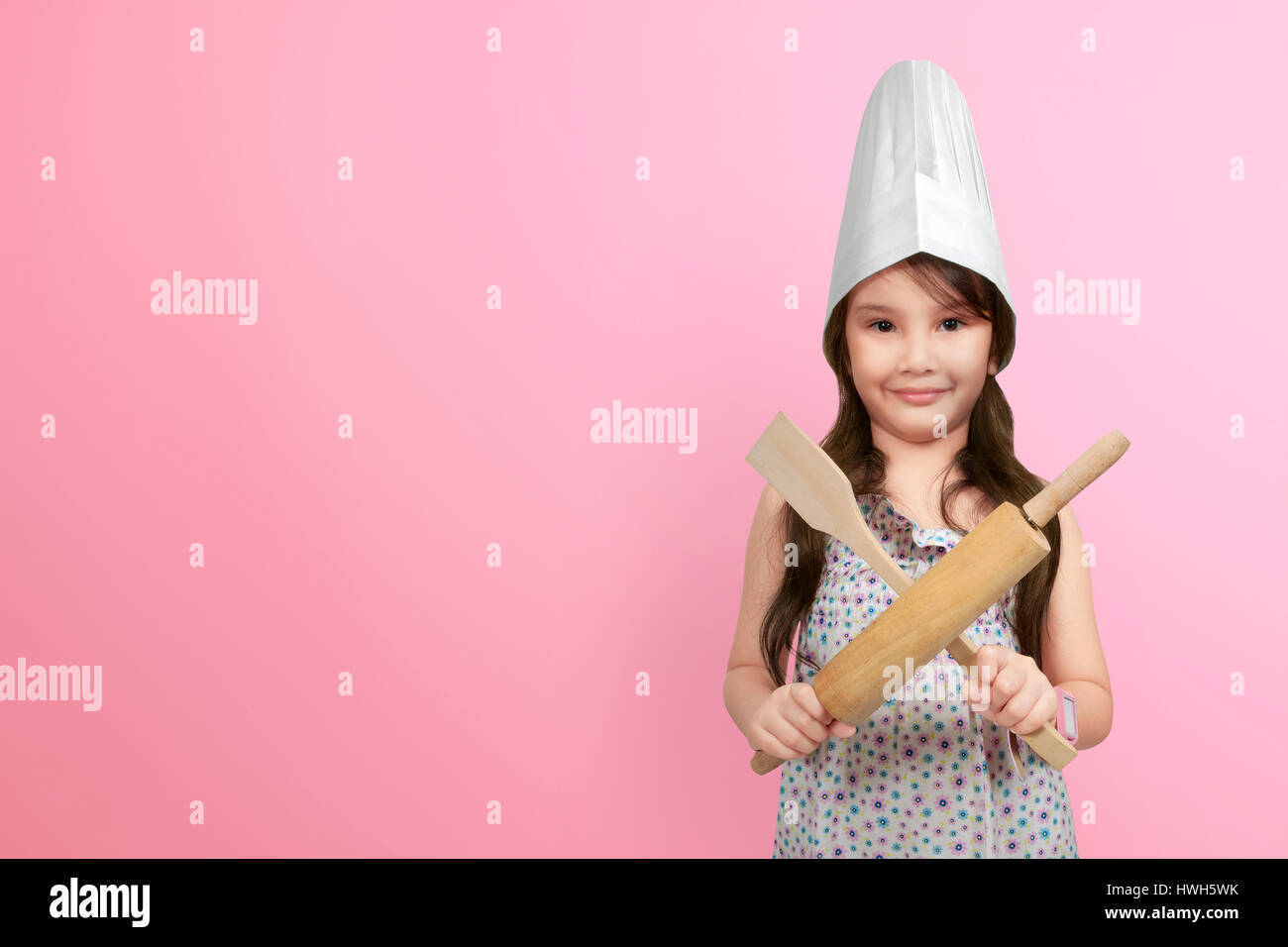 Smiling asian little girl in chef hat holding wooden cooking utensils over pink background Stock Photo