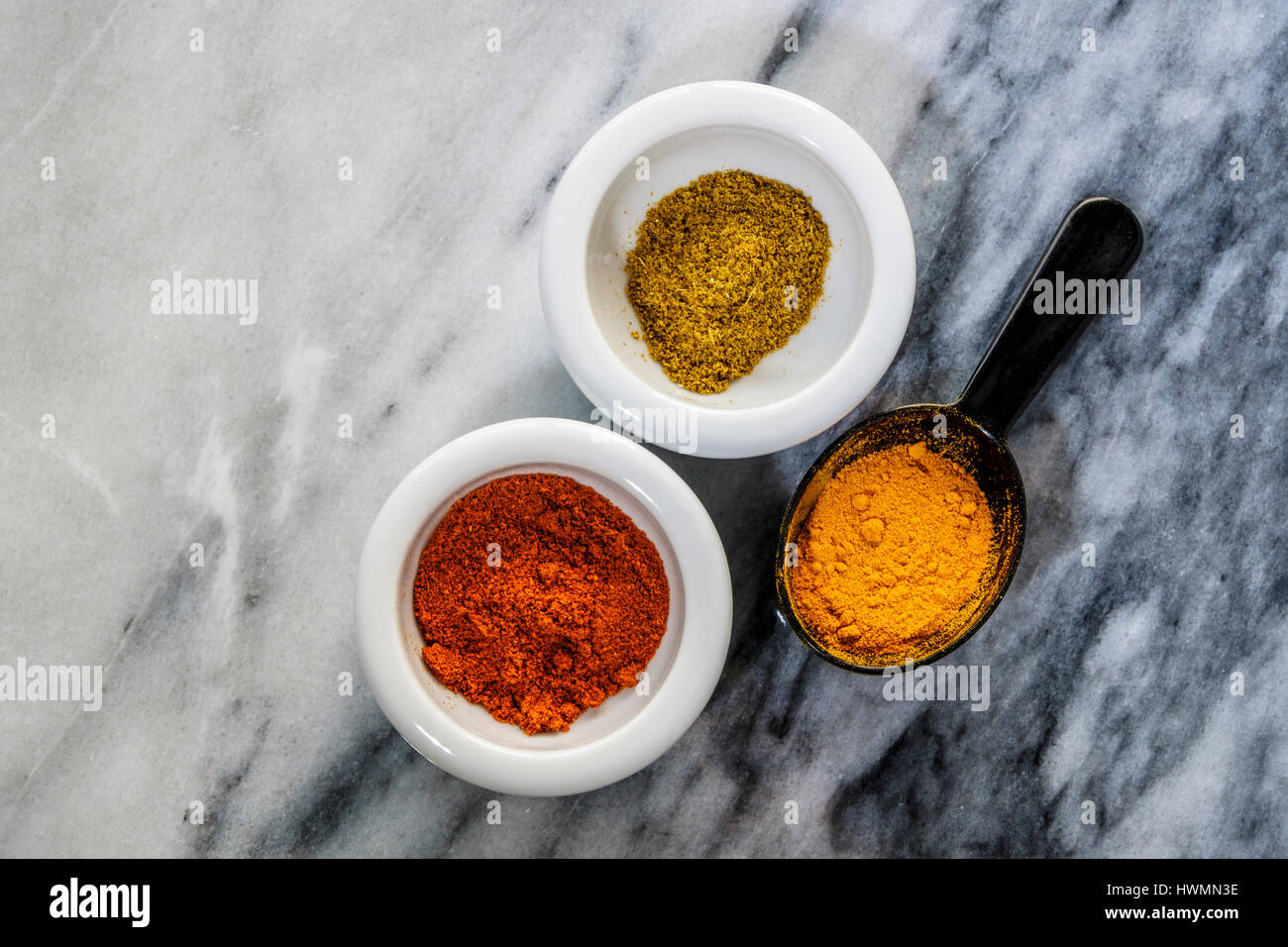 Vibrant colors of spices in small white dishes and black measuring spoon lay flat on marble. Turmeric, cumin and paprika. Stock Photo