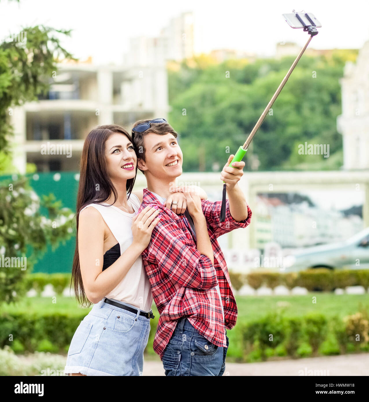 happy young tourists couple taking a selfie with smartphone on the monopod in city. The man is holding the stick and shooting looking at phone with ha Stock Photo