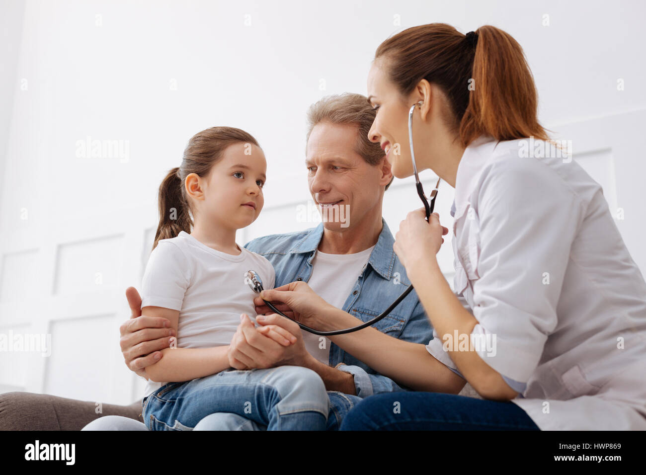 What are your complains. Gentle trained professional pediatrician using her stethoscope checking her little patients lungs while her dad holding her o Stock Photo