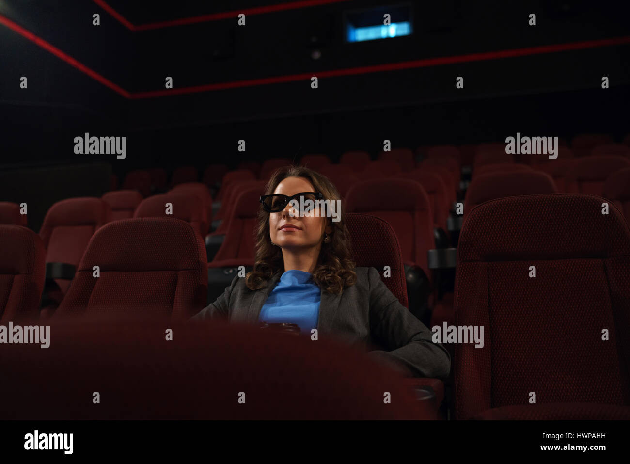 Young woman watches movie 3D at the cinema and drinks coffee, looking at the screen, alone in an empty room, overall plan Stock Photo