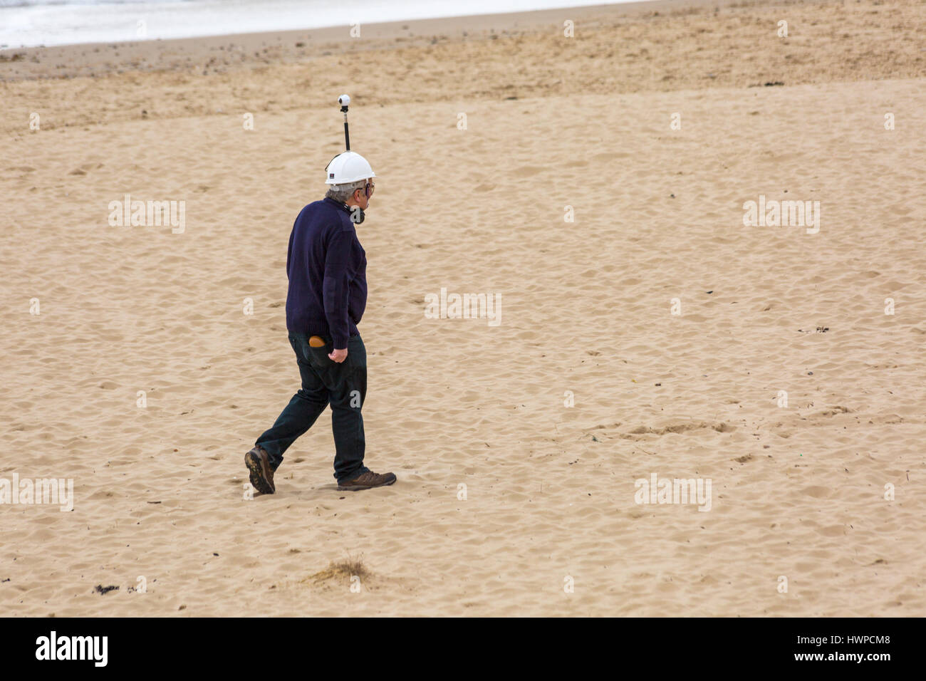 Man walking along beach wearing helmet with pole and camera at Sandbanks, Poole, Dorset in March Stock Photo