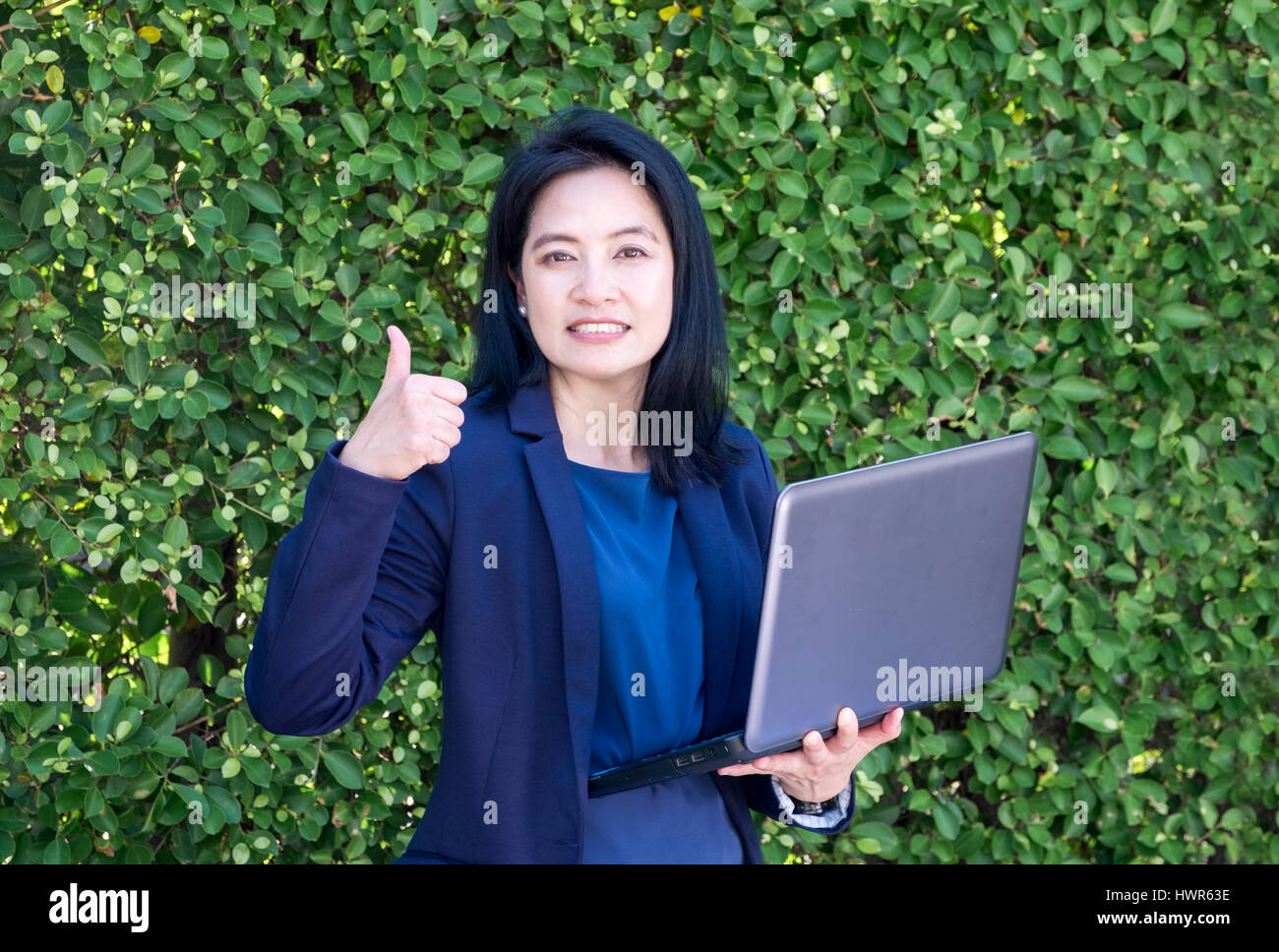 businesswoman thumps up and holding laptop computer at green leaf wall outside office,Good success plan. Stock Photo