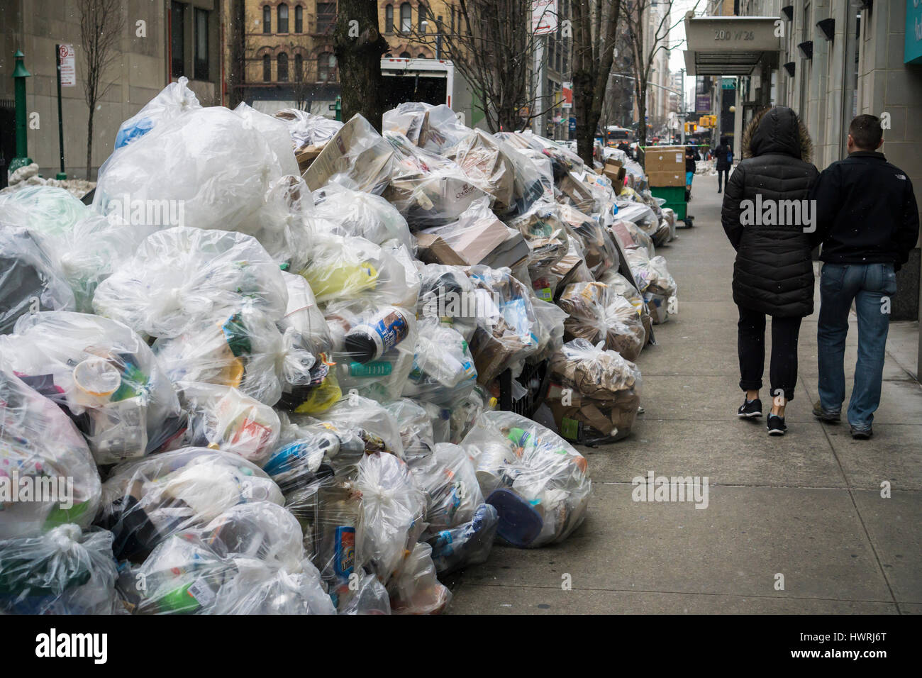 Pedestrians walk past bags of recyclable trash awaiting pickup in the