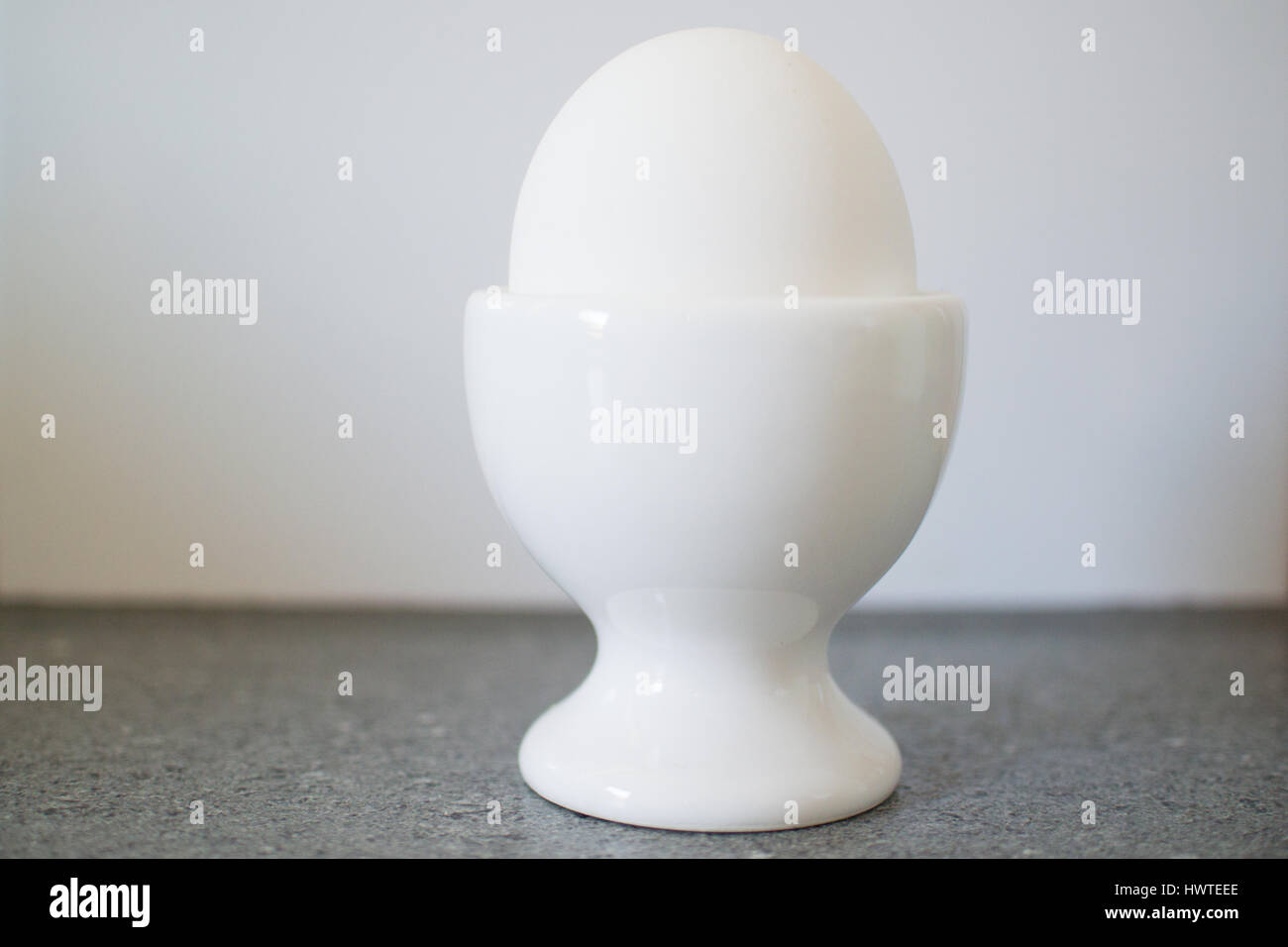 Hard boiled white egg in white egg cup, in front of white wall Stock