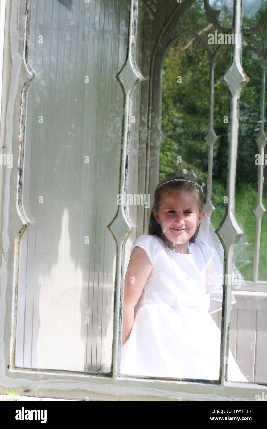 Girl in a first communion dress looking through a glass window Stock Photo