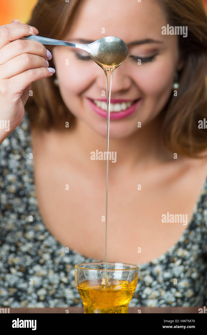 Young woman facing camera holding spoon with honey dripping into small glass on table Stock Photo