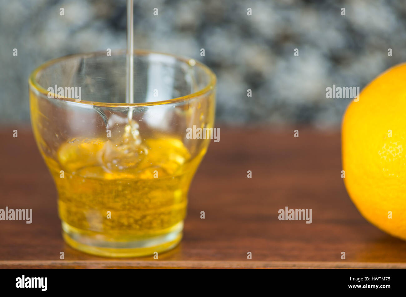 Close up small glass sitting on wooden desk with honey falling into it from above, lemon on the side Stock Photo