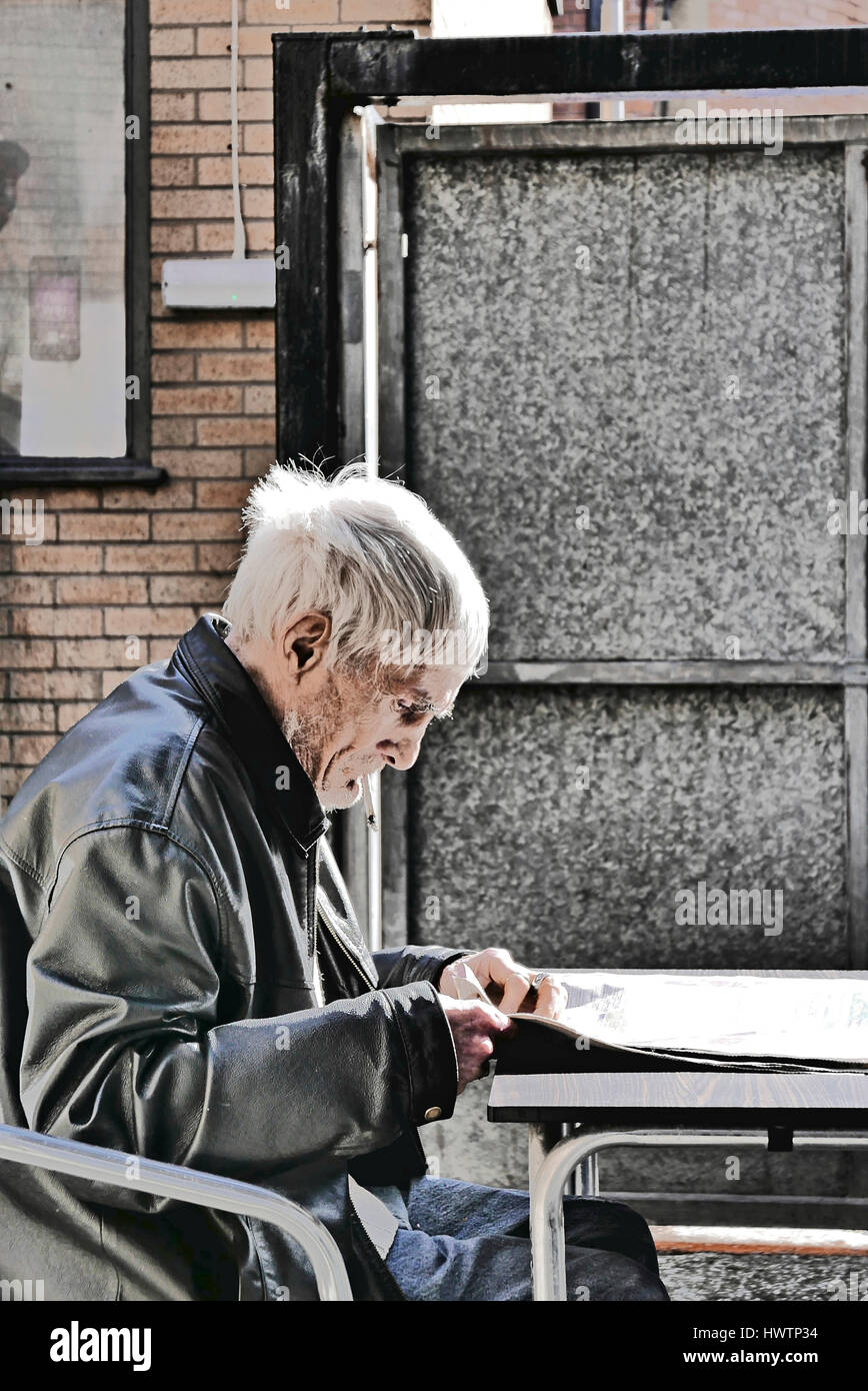 Senior man in leather jacket sat outside smoking and reading a newspaper Stock Photo