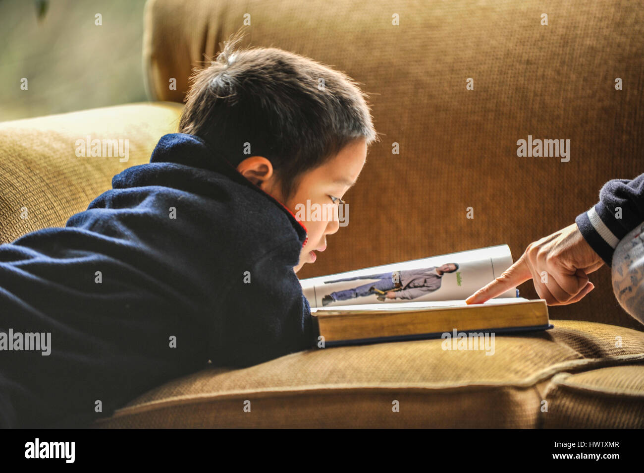 A young boy on a couch reading a big picture book.  A mother's finger pointing.  Close up, natural light, horizontal, non-posing Stock Photo