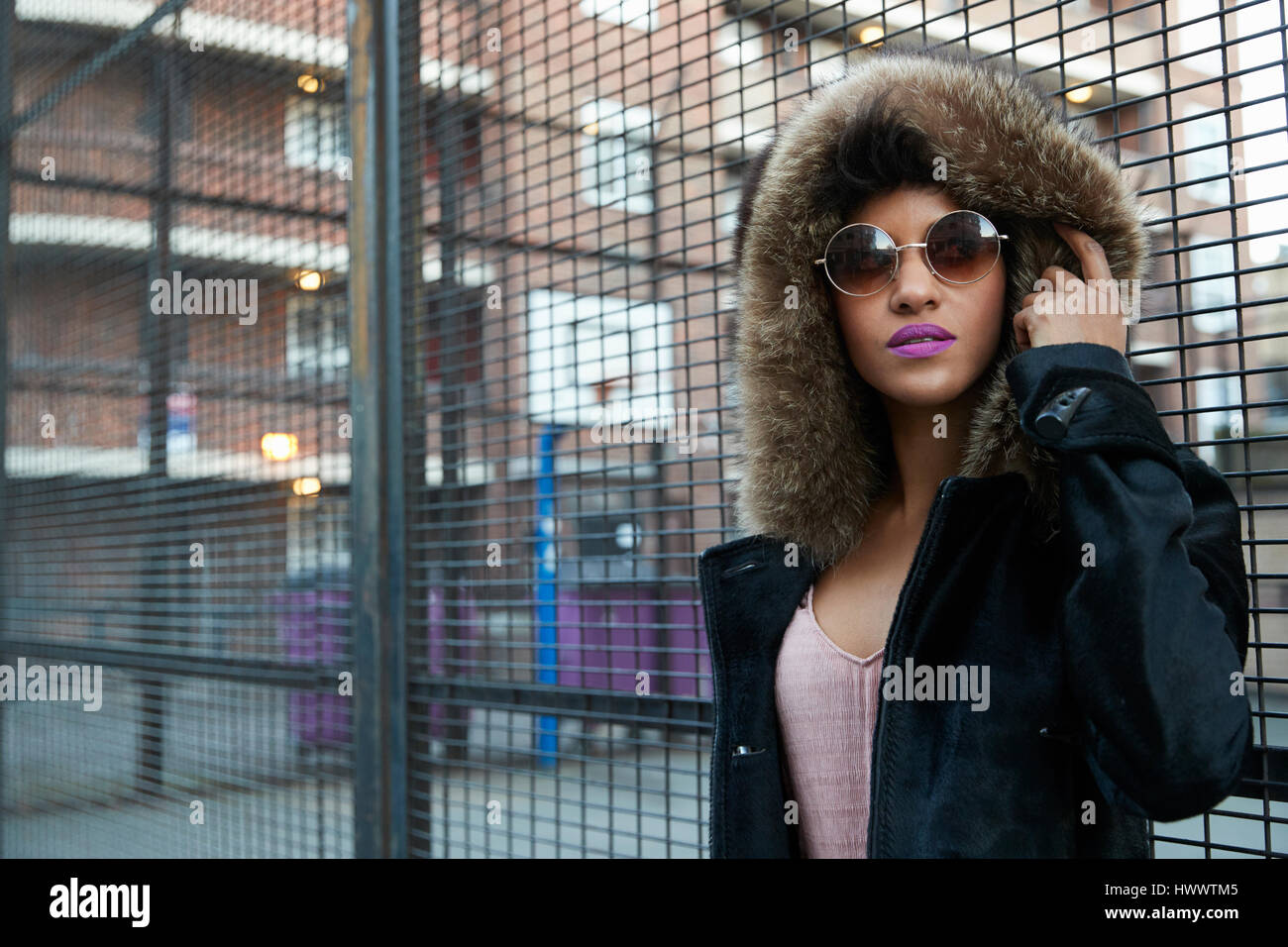 Young woman in fur-lined hood standing by fence, horizontal Stock Photo