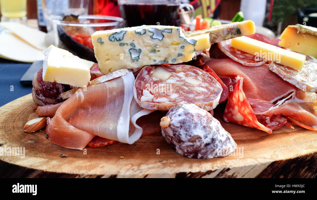 Cuts of blue, cow, goat cheese, pork ham, smoked meats, salami, fat sausages served on a piece of wood for lunch in apres ski chalet bistro . Stock Photo