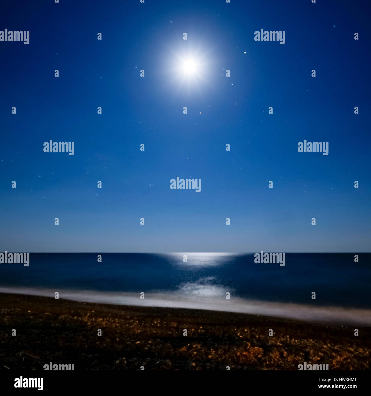 bright moon surrounded with stars reflected in the water at the beach at night Stock Photo