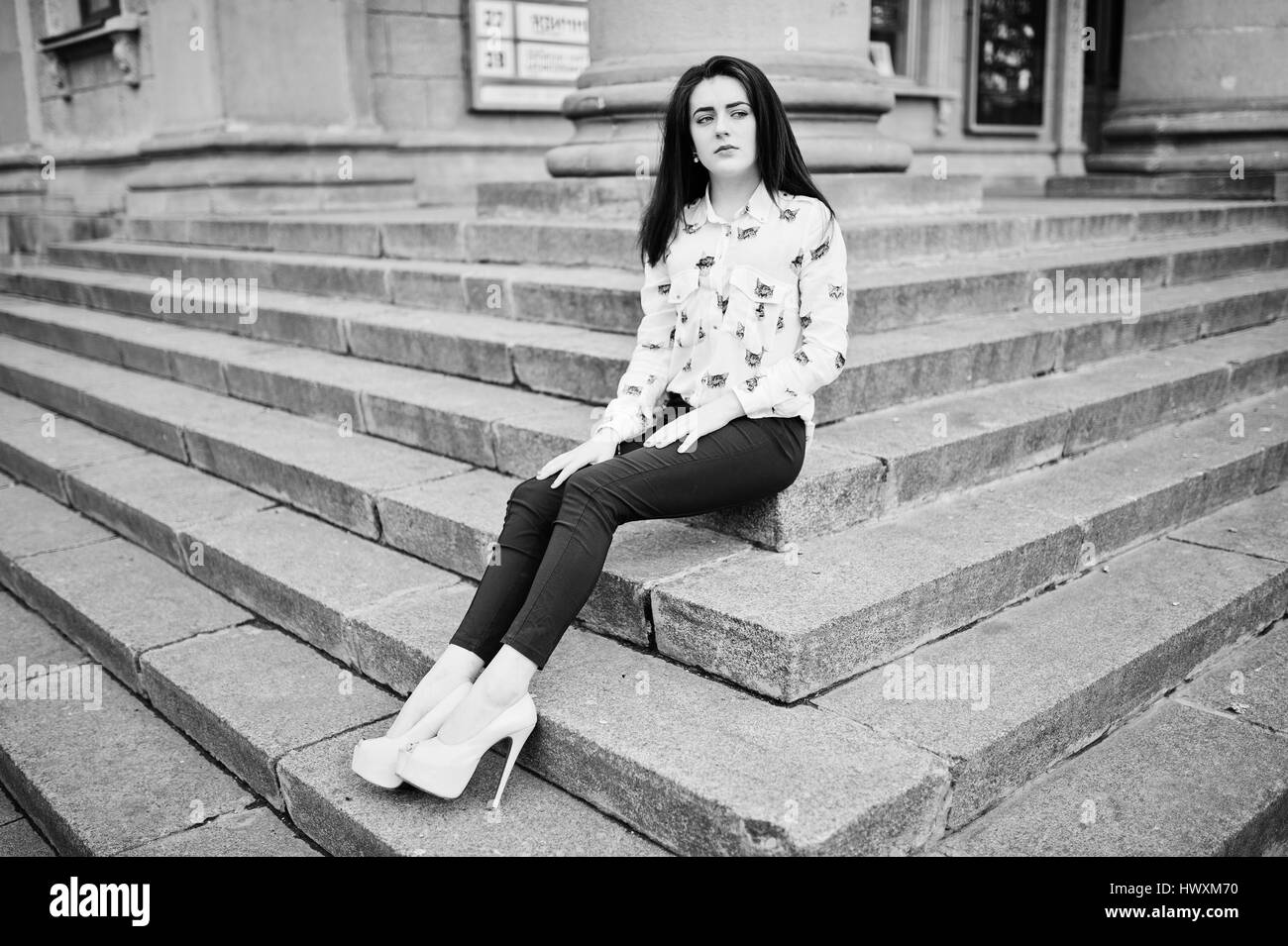 Young stylish brunette girl on shirt, pants and high heels shoes, posed at stone stairs. Street fashion model concept. Black and white photo. Stock Photo