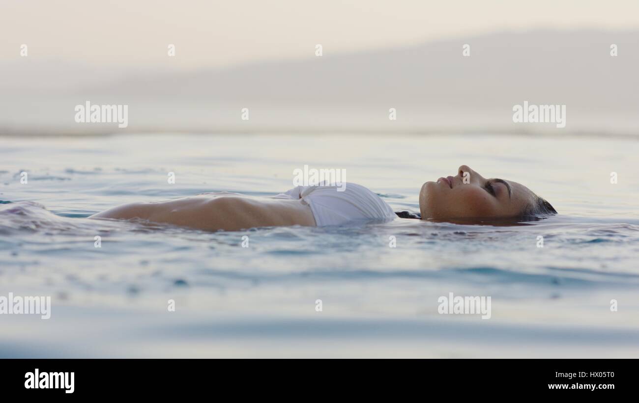 Profile of woman in bikini floating in infinity pool overlooking horizon and remote landscape Stock Photo