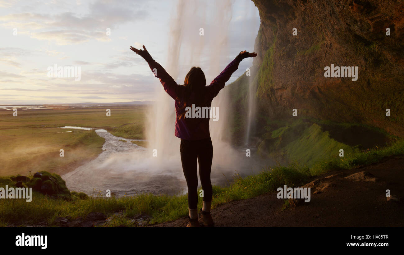 Rear view silhouette of woman with outstretched arms standing behind waterfall overlooking remote landscape Stock Photo