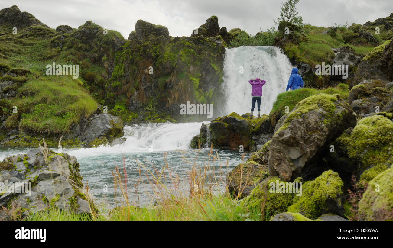 Couple standing on mossy boulders admiring waterfall river in rocky remote landscape Stock Photo