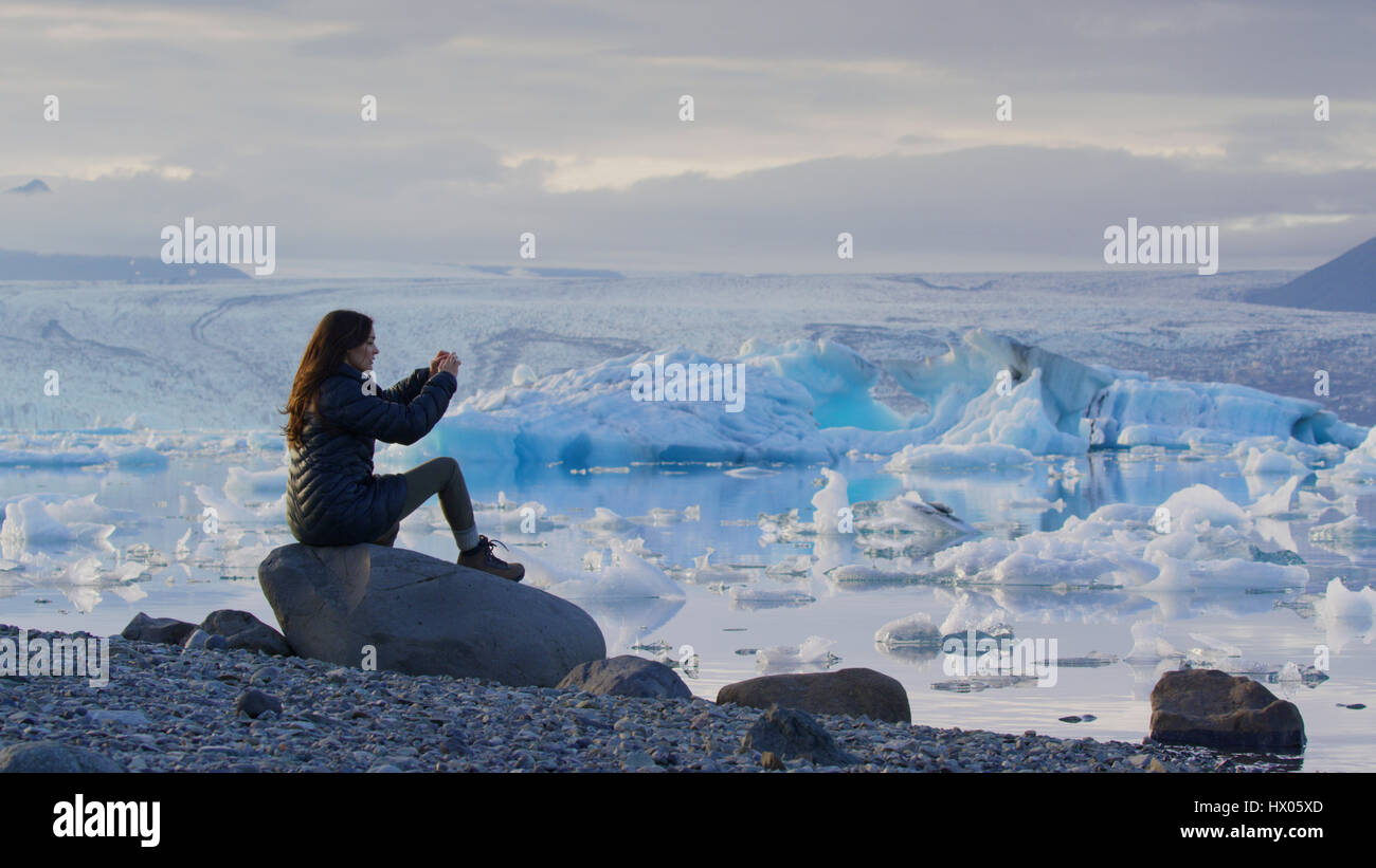 Profile view of woman photographing remote landscape and frozen glaciers in still ocean under cloudy sky Stock Photo