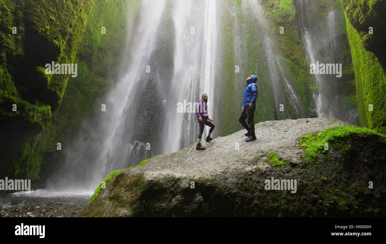 Low angle view of couple standing on mossy boulder admiring waterfall in remote landscape Stock Photo