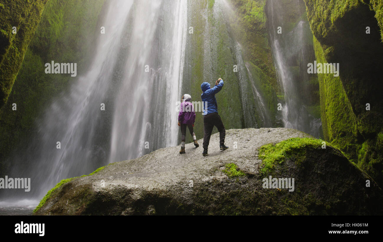 Low angle view of couple taking pictures with smartphone standing on mossy boulder near waterfall in remote landscape Stock Photo