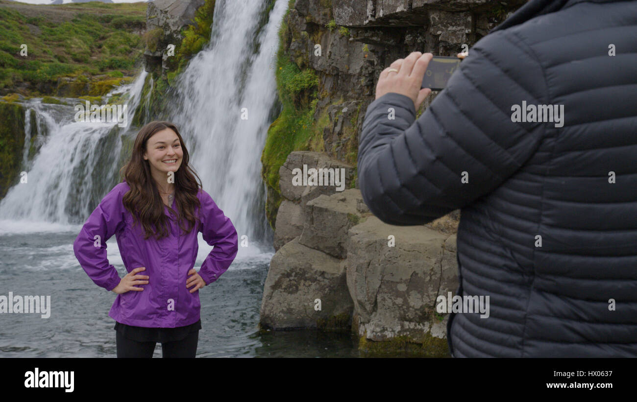 Man photographing smiling girlfriend posing near waterfall over remote rock cliffs Stock Photo