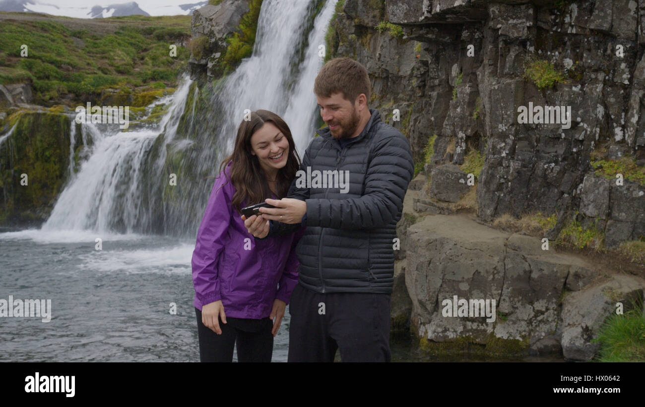 Boyfriend and girlfriend using cell phone near waterfall over remote rock cliffs Stock Photo