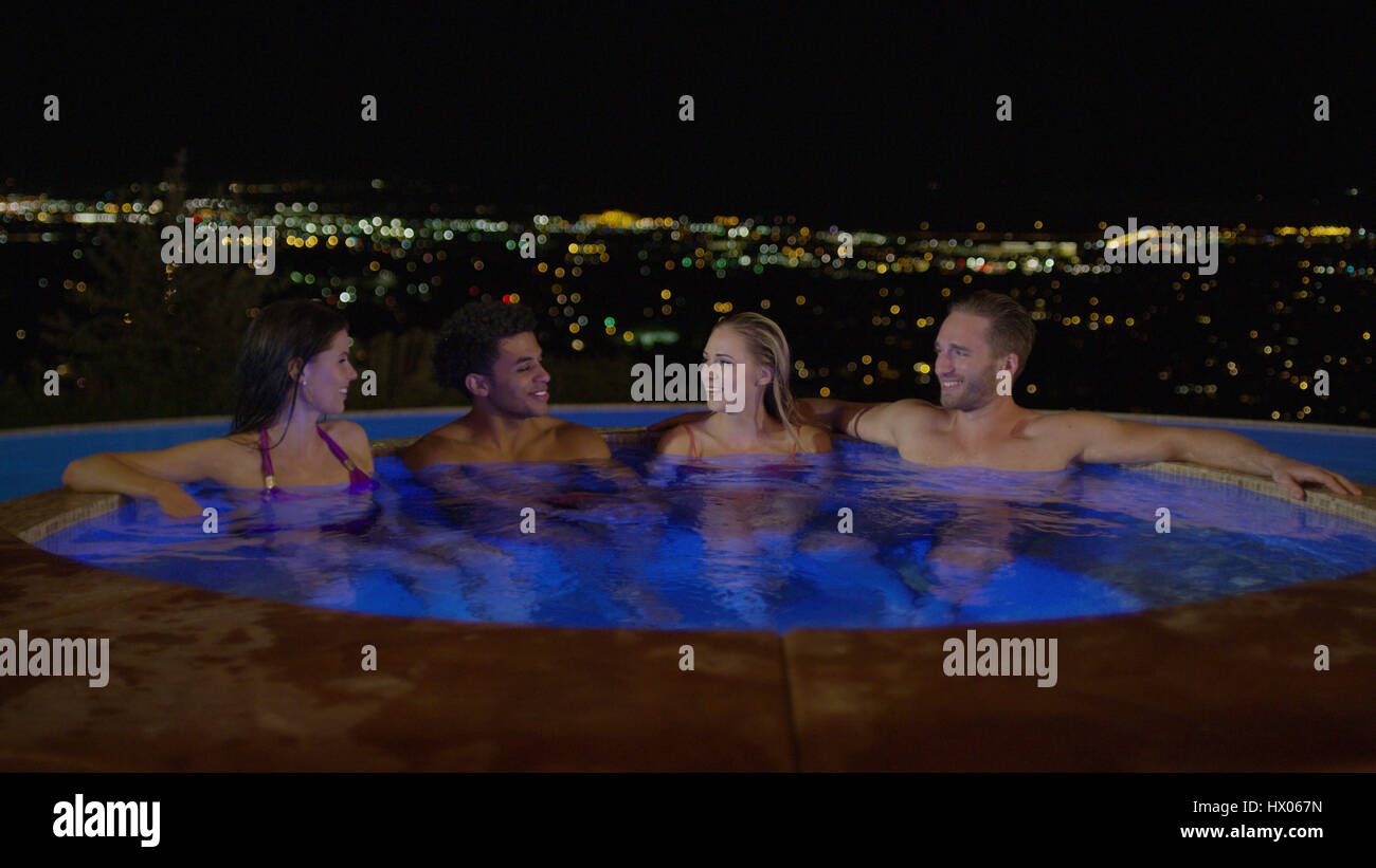 Smiling couples sitting in illuminated hot tub overlooking cityscape at night Stock Photo