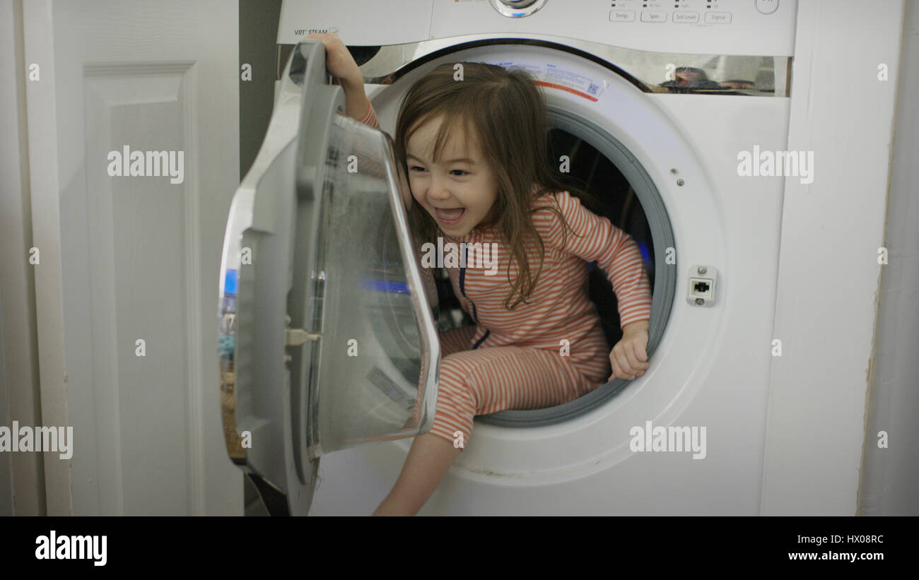 Laughing playful girl climbing in dryer appliance in laundry room Stock Photo