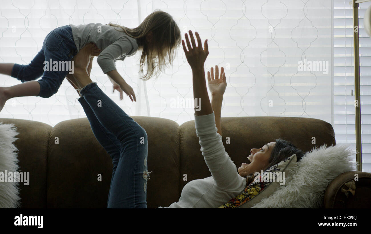 Profile of laughing playful mother lifting daughter in mid-air and playing on sofa Stock Photo