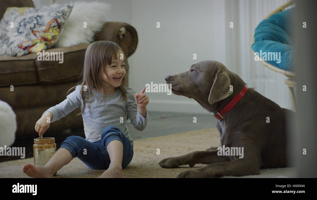 Naughty girl feeding pet dog peanut butter on living room floor Stock Photo