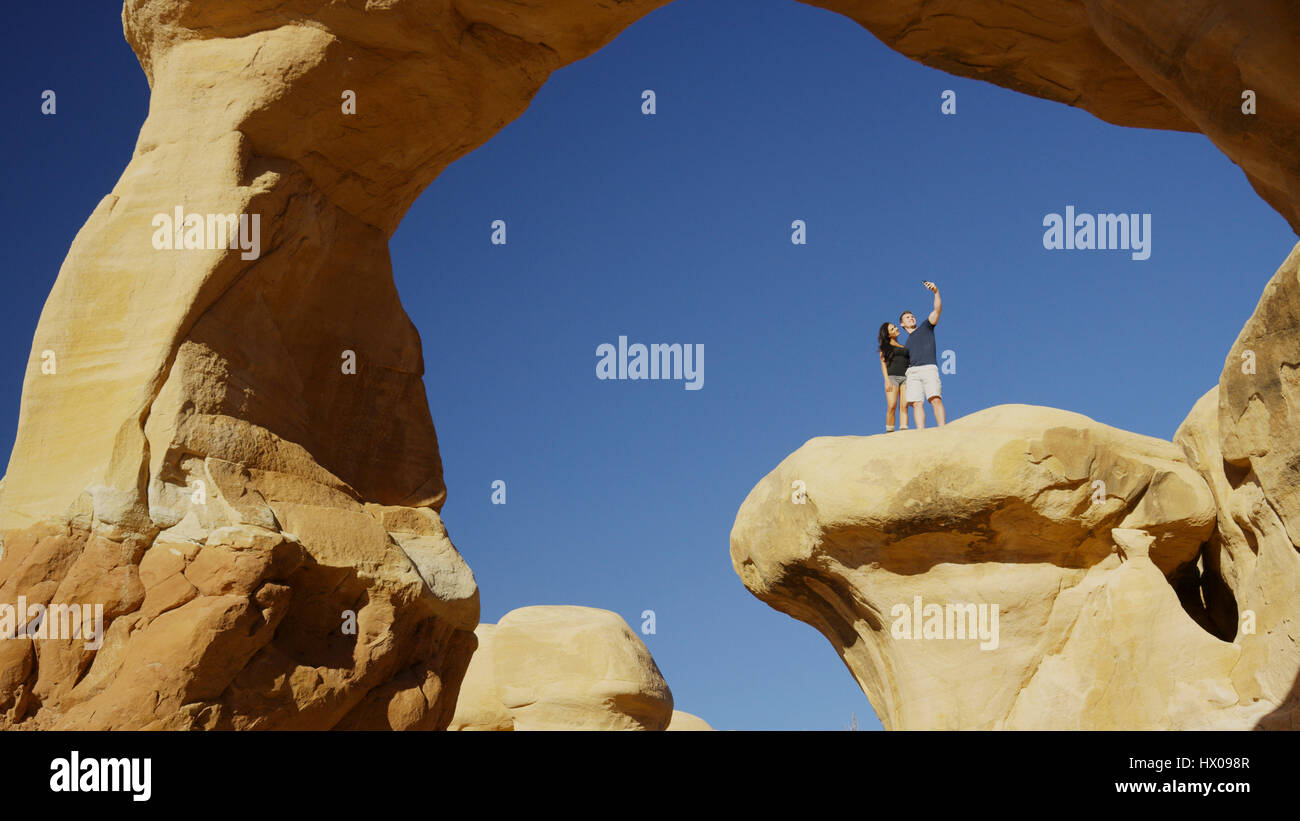 Low angle view of boyfriend and girlfriend taking selfie with smartphone on scenic rock formations under clear blue sky Stock Photo